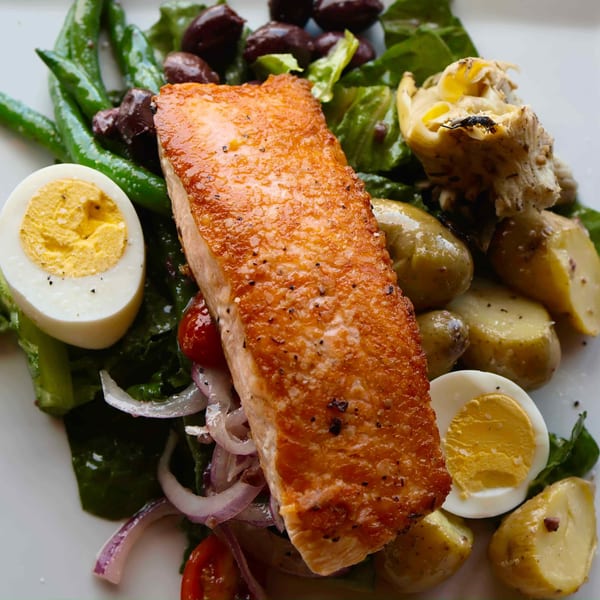 Seared salmon with green beans, olives, potatoes, hard-boiled eggs, and red onion on a white plate.