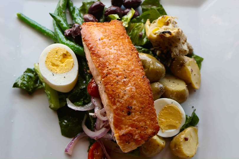 Seared salmon with green beans, olives, potatoes, hard-boiled eggs, and red onion on a white plate.